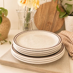 May include: Four speckled ceramic plates with brown rims, stacked on a beige book. A bouquet of yellow and white flowers in a clear vase is in the background, along with a wooden chopping board and a potted plant.