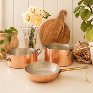 Puede incluir: Conjunto de utensilios de cocina de cobre, que incluye dos cacerolas y una sartén, con asas de latón. También se ven una tabla de cortar de madera y un jarrón con flores. La escena tiene lugar en una cocina luminosa.