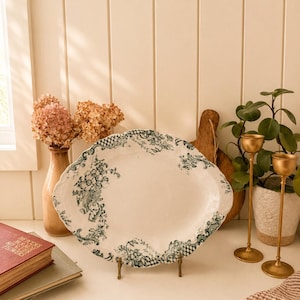 May include: An oval ceramic platter featuring a white base with a dark green transferware pattern of floral motifs and lattice borders along the scalloped edges. The platter is displayed upright on a small metal stand, showcasing its detailed design.