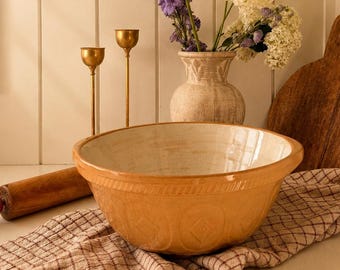 Puede incluir: Un cuenco grande y redondo de cerámica esmaltada en amarillo con interior color crema y patrones de diamantes en relieve en el exterior, sobre un paño a cuadros beige y marrón junto a un rodillo de madera y una tabla de cortar de madera.