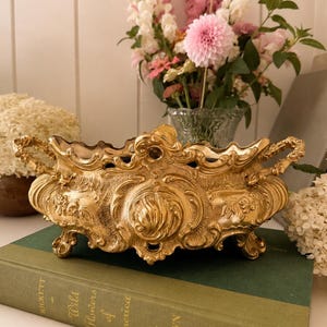 Peut inclure: Un bol décoratif en métal doré orné de volutes complexes, de motifs floraux en relief et de deux poignées latérales. Le bol est soutenu par quatre petits pieds et repose sur un épais livre à couverture rigide vert intitulé Wild Flowers of America.