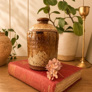 Peut inclure: Un pot en céramique artisanal avec un couvercle, aux tons bruns et beiges. Il repose sur un livre rouge vintage. Un petit bouquet de fleurs séchées et un bougeoir doré sont également présents.