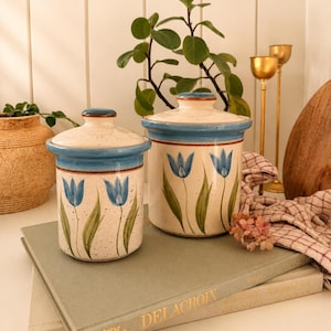 Könnte beinhalten: Zwei Keramikbehälter mit Deckeln. Die Behälter sind cremefarben mit blauen Tulpenmotiven und blauen Rändern. Sie stehen auf Büchern. Eine Pflanze und goldene Kerzenhalter sind im Hintergrund.