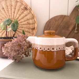 May include: A retro ceramic teapot in shades of brown and white, featuring a speckled glaze. The teapot has a handle, spout, and lid. It rests on books. A woven basket and wooden board are in the background, creating a vintage aesthetic.