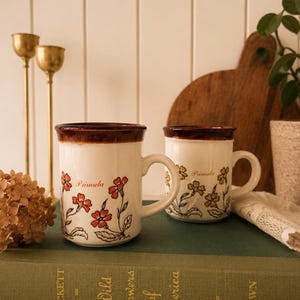 May include: Two ceramic mugs with a cream-colored base and a dark brown rim. The left mug features red floral illustrations and the word Primula, while the right mug displays yellow floral illustrations and the word Primula in a script font.