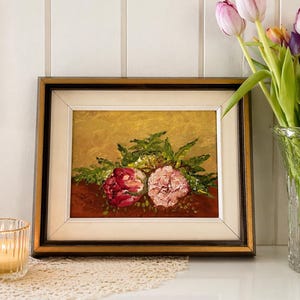 May include: Framed floral oil painting featuring two large flowers in shades of red and pink, surrounded by green foliage. The painting has a gold background and a dark brown frame. A vase of tulips sits nearby.