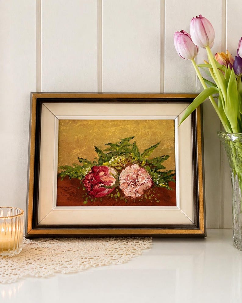 May include: Framed floral oil painting featuring two large flowers in shades of red and pink, surrounded by green foliage. The painting has a gold background and a dark brown frame. A vase of tulips sits nearby.