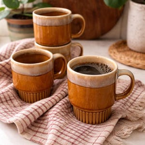 Könnte beinhalten: Vier Keramikbecher in Braun- und Cremetönen, einer mit Kaffee gefüllt. Sie stehen auf einem rosa-weiß karierten Tuch. Die Becher haben einen gerippten Boden und einen abgerundeten Griff. Ein Holzbrett und Pflanzen sind im Hintergrund zu sehen.