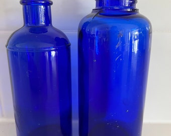 May include: Two cobalt blue glass bottles of different sizes. The bottle on the left is slender, while the one on the right is wider and taller. Both bottles have a threaded neck.