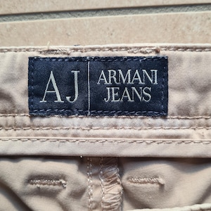 May include: Close-up of beige-colored pants with a dark blue label that reads "AJ ARMANI JEANS" in white lettering. The pants have visible stitching and a metal button. The image is taken on a light brown surface.