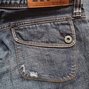 May include: Close-up of blue denim jeans with a brown leather label that reads "Replay" and "Famous Quality". The jeans have a unique pocket design with a button closure and distressed detailing.