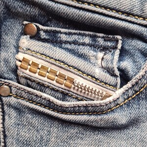 May include: Close-up of a denim jeans pocket with a decorative zipper. The pocket features a silver zipper with gold-colored teeth and is stitched with yellow thread. The denim has a faded, worn appearance.