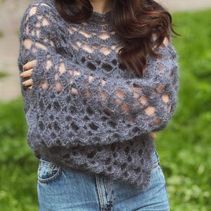 Könnte beinhalten: Ein grauer Häkelpullover mit offenem Gewebe und langen Ärmeln. Der Pullover hat einen runden Ausschnitt und wird mit einer blauen Jeans kombiniert. Das Häkelmuster erzeugt eine zarte, luftige Textur, ideal zum Überziehen.