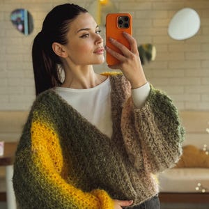 May include: A woman wearing a chunky knit sweater with a gradient of yellow, green, and brown hues. The sweater has a V-neck and long, voluminous sleeves. She is holding an orange phone and taking a selfie.