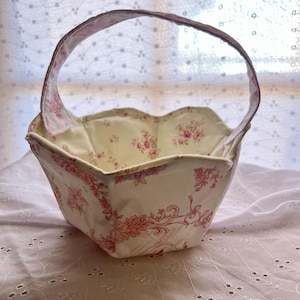 May include: A fabric basket with a handle, featuring a floral pattern in pink and white. The interior has a light yellow fabric with pink roses. The basket's exterior showcases a detailed pink floral design on a white background.