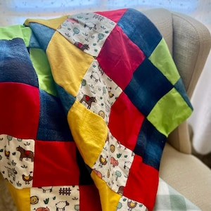 May include: A patchwork quilt with a farm animal theme. The quilt features squares of red, yellow, blue, and green fabric. Some squares have printed images of sheep, cows, chickens, and horses. The quilt is draped over a chair.
