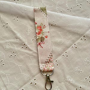 May include: A floral patterned lanyard with a silver-colored clasp. The fabric features a light pink background with a repeating pattern of small white dots and red roses. The lanyard is approximately 1 inch wide.