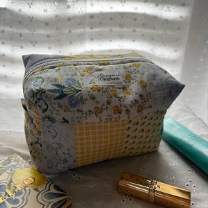 May include: A quilted cosmetic bag with a patchwork design featuring floral and yellow gingham patterns. The bag has a gray zipper and a label that reads "Gossamer Gingham." A gold lipstick case, a blue and white patterned book, and a tube of lotion are also visible.