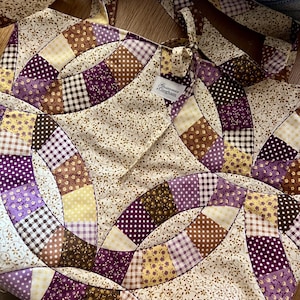 May include: A fabric tote bag featuring a patchwork design in shades of purple, brown, and cream. The pattern consists of interlocking curved segments with floral, polka dot, and gingham prints. A white label reads Gossamer Gingham.