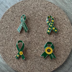 May include: Four green ribbon pins with various designs. One pin has the text "IN DISABILITY VISIBLE". Other pins feature yellow flowers and a sunflower. The pins are on a round, cork surface.