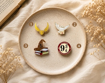May include: Four enamel pins on a speckled plate: a gold winged ball, a white owl carrying an envelope, a brown wizard hat resting on a stack of books, and a circular red and white sign with the text Platform 9 3/4 Hogwarts Express.