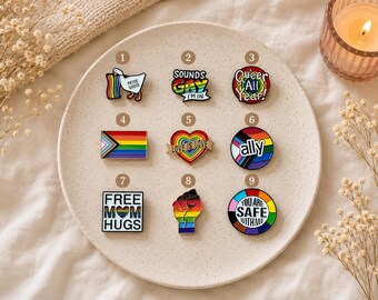 May include: Nine enamel pins arranged on a plate. 1: Goose with rainbow flag and text PRIDE GOOSE. 2: Text SOUNDS GAY I'M IN. 3: Circular pin with rainbow and text QUEER ALL YEAR. 4: Progress Pride flag. 5: Heart with rainbow and text LOVE IS LOVE. 6: Circular pin with text ally. 7: Square pin with text FREE MOM HUGS. 8: Rainbow fist. 9: Circular pin with text YOU ARE SAFE WITH ME.