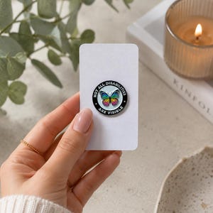Puede incluir: Un pin esmaltado redondo que presenta una mariposa arcoíris colorida en el centro sobre un fondo azul claro. El borde negro contiene el texto blanco NOT ALL DISABILITIES ARE VISIBLE. El pin está sujeto a una tarjeta de presentación blanca rectangular.