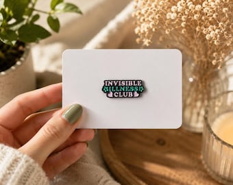 May include: A rectangular enamel pin with a black border and a light pink background. The text reads INVISIBLE ILLNESS CLUB in bold, dark-colored letters, with the word ILLNESS in teal. Two small light pink hearts flank the text on each side.
