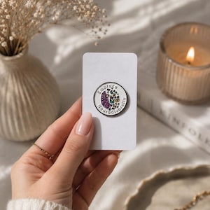 Peut inclure: Une broche émaillée circulaire avec un fond blanc et un design divisé. Un côté présente une illustration de cerveau violet, tandis que l'autre côté affiche une collection de papillons colorés. Le texte Embrace Neurodiversity entoure le bord.