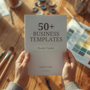 May include: A person holding a white book titled "50+ Business Templates" with "Studio Toolkit" and "System Tools" printed on the cover. The book is on a wooden surface with art supplies and colour swatches.