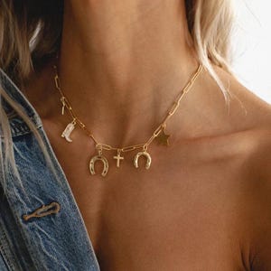 May include: Gold-colored necklace with a paperclip chain design. The necklace features charms including a cowboy boot, a star, a cross, and two horseshoes. The necklace is worn around the neck.