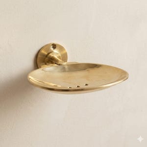 May include: A wall-mounted brass soap dish with a circular backplate and an oval-shaped dish. The dish has a slight curve and three drainage holes. The fixture is made of shiny, polished brass and is mounted on a light-colored wall.