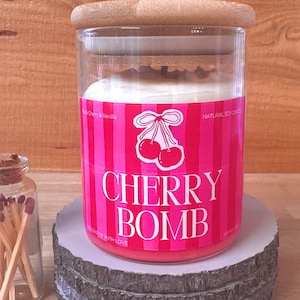 May include: A clear glass candle jar with a light wood lid, filled with white wax. The label is bright pink with vertical stripes, a cherry illustration, and the text "CHERRY BOMB", "Black Cherry & Vanilla", and "NATURAL SOY CANDLE".