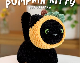 May include: A black crocheted cat plushie wearing a yellow pumpkin-shaped hood with a green stem and leaf detail. The text PUMPKIN KITTY PDF PATTERN is displayed in white above the cat. The cat has bright green eyes and a small white nose.