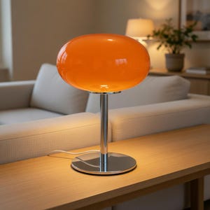 Peut inclure: Lampe de table en verre orange avec un abat-jour arrondi et une base chromée. La lampe est posée sur une table en bois. L'abat-jour est d'une couleur orange vif.