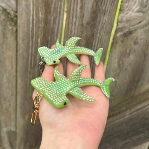 May include: Two green hammerhead shark keychains with white polka dots. The keychains have a gold clasp and silver chain. The shark keychains are made of a smooth material and are a fun accessory.