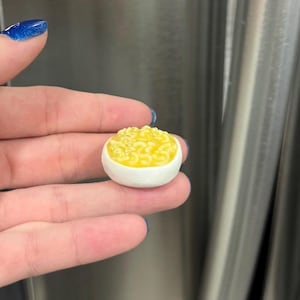 May include: A miniature bowl of macaroni and cheese. The bowl is white, and the macaroni is yellow. The image is a close-up shot, highlighting the details of the food. The bowl is held between two fingers.