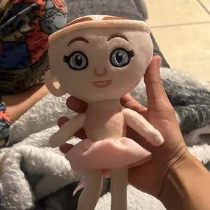 May include: A plush doll shaped like a teacup with a face, arms, and legs. The doll has a light pink tutu, ballet slippers, and a matching headband. The eyes are blue, and the doll is held in a person's hand.