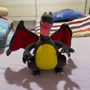 May include: A plush Charizard toy in a standing pose. The toy features a black body, yellow belly, red wings, and a pink tongue. The toy has white claws and is set against a soft background.