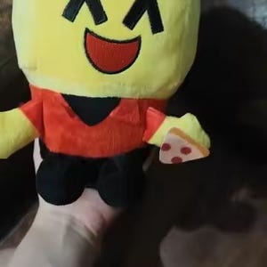 May include: A plush toy in the shape of a character with a yellow head, red hat and shirt, black accents, and black shoes. The character is holding a slice of pizza. The toy has a smiling face.