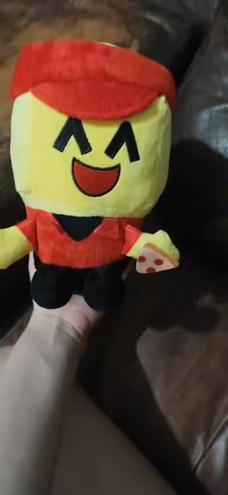 May include: A plush toy in the shape of a character with a yellow head, red hat and shirt, black accents, and black shoes. The character is holding a slice of pizza. The toy has a smiling face.