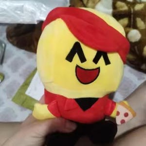 May include: A plush toy character with a yellow head, red hair and shirt, black pants, and a slice of pizza. The character has a smiling face with black eyes and a red mouth. The toy is held by a person.