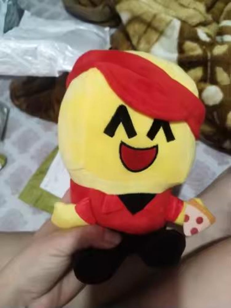May include: A plush toy character with a yellow head, red hair and shirt, black pants, and a slice of pizza. The character has a smiling face with black eyes and a red mouth. The toy is held by a person.