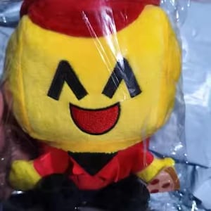 May include: A plush toy in a clear plastic bag. The toy has a yellow head with black eyes and a red smile. It wears a red hat and shirt with black pants. A small piece of pizza is in its hand.
