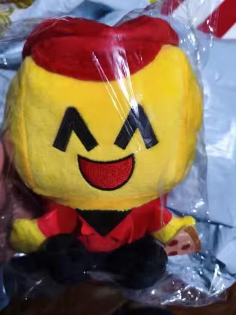 May include: A plush toy in a clear plastic bag. The toy has a yellow head with black eyes and a red smile. It wears a red hat and shirt with black pants. A small piece of pizza is in its hand.
