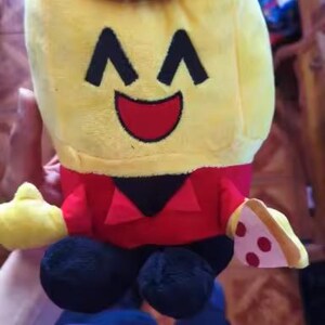May include: A plush toy shaped like a smiling yellow character wearing a red hat and shirt. The character has black eyes and a red mouth. It holds a slice of pizza and has black feet.