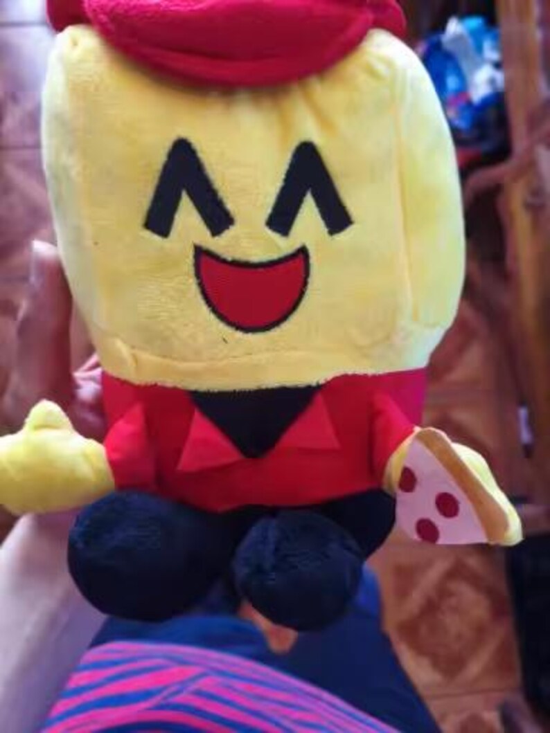 May include: A plush toy shaped like a smiling yellow character wearing a red hat and shirt. The character has black eyes and a red mouth. It holds a slice of pizza and has black feet.