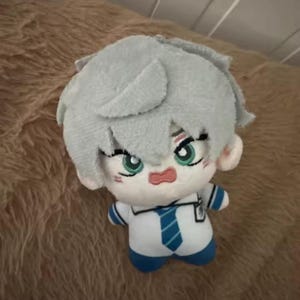 May include: A plush doll with light blue hair and green eyes. The doll is wearing a white shirt with a blue tie and blue shorts. The doll has a surprised expression and is sitting on a brown surface.