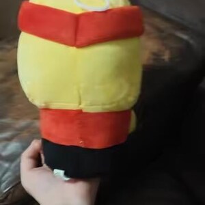 May include: A plush toy with a yellow head and body, a red band around the head and waist, and a black base. A white ring is on top of the head. The toy is held in a person's hand.
