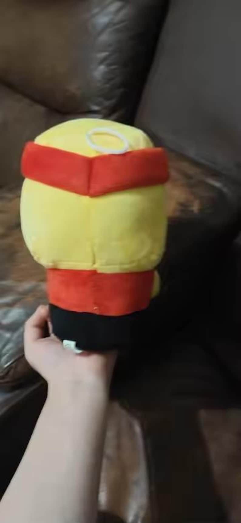 May include: A plush toy with a yellow head and body, a red band around the head and waist, and a black base. A white ring is on top of the head. The toy is held in a person's hand.